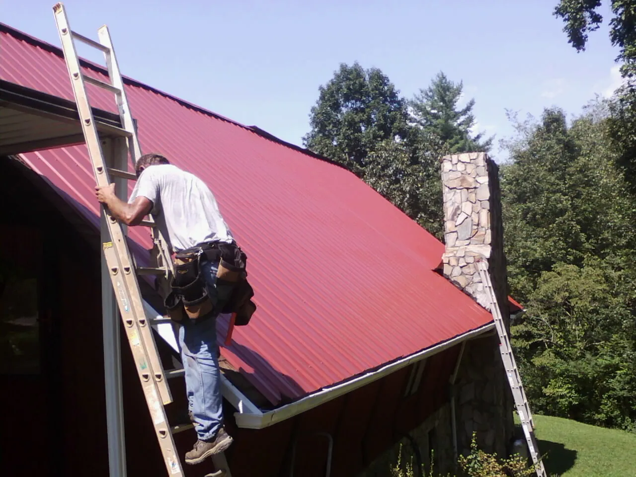 Roofer on a ladder beside a red metal roof and stone chimney — Soffit & Fascia Repair by Roofing Brothers in Williamsport